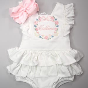 Puede incluir: Romper blanco para bebé con capas con volantes y bordado floral. Se adjunta un gran lazo rosa. El romper presenta el nombre "Collins" bordado dentro de una corona floral. El diseño incluye detalles florales rosas y azules.