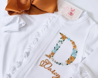 May include: A white long-sleeve romper with ruffled trim, featuring an embroidered floral letter D and the name Darcy in gold thread. A large, solid burnt orange hair bow sits above the romper, accompanied by a tag labeled fox bloom.