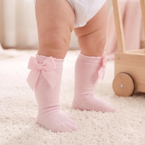 May include: A pair of light pink ribbed knee-high socks featuring a decorative satin bow attached to the outer side of each cuff. The socks are made of a soft knit fabric and extend to the mid-calf area, providing a snug fit around the lower legs.