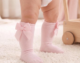 May include: A pair of light pink ribbed knee-high socks featuring a decorative satin bow attached to the outer side of each cuff. The socks are made of a soft knit fabric and extend to the mid-calf area, providing a snug fit around the lower legs.