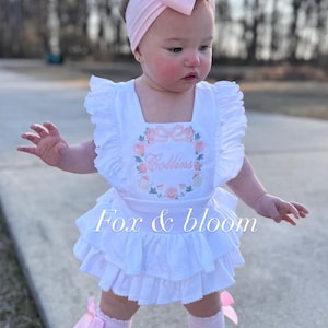 May include: A white baby dress with a floral design and the name "Collins" embroidered on the front. The dress has ruffled sleeves and a tiered skirt. The outfit includes a matching pink headband, socks, and shoes. The text "Fox & bloom" is also visible.