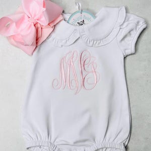 Könnte beinhalten: Weißer Baby-Strampler mit Peter Pan-Kragen und rosa gestickten Initialen. Eine große rosa Schleife ist befestigt. Der Strampler hat einen Rüschenkragen und kurze Ärmel. Ein Etikett mit einem Logo ist angebracht.