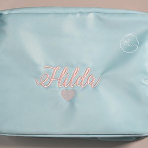 May include: A light blue cosmetic bag with the name "Hilda" embroidered in pink script, along with a pink heart. The bag has a rectangular shape and a gold zipper. The bag is made of a smooth, possibly nylon, material.