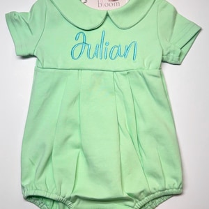 Könnte beinhalten: Ein hellgrüner Baby-Strampler mit einem Bubikragen und kurzen Ärmeln. Der Name "Julian" ist in blauer Schrift auf die Brust gestickt. Ein Etikett mit einem Fuchlogo ist am Kragen befestigt. Der Strampler hat einen gerafften, ballonartigen Unterteil.