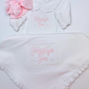 Puede incluir: Una camisa blanca de manga larga con cuello y puños con volantes, junto a una manta blanca a juego con borde de volantes. Ambos artículos tienen bordado el nombre Jazzlyn Jae en hilo rosa dentro de un marco rectangular festoneado.