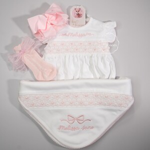 Puede incluir: Un conjunto de ropa de bebé blanco con detalles en rosa. El conjunto incluye un vestido, un babero, calcetines y un lazo grande. El vestido y el babero presentan detalles bordados y el nombre "Melissa Jane". La etiqueta de la marca dice "fox & bloom".