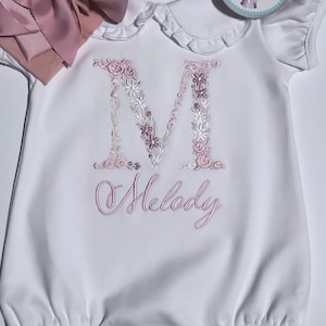 May include: White baby onesie with a pink floral embroidered letter M and the name Melody embroidered below. The onesie has a ruffled collar and a pink bow headband.