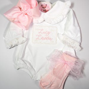 Op de afbeelding: Een witte romper met lange mouwen, een kraag met ruches, elastische boorden en de naam Lucy Lavon geborduurd in roze schrijfletters. De set bevat een grote roze haarstrik en een paar lichtroze sokjes versierd met een transparante roze strik.