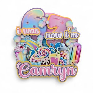 Puede incluir: Adorno para pastel colorido con el texto "i was now i'm" y el nombre "Camryn". El diseño incluye ilustraciones de dulces como helados, cupcakes, piruletas y un tarro de malvaviscos. La paleta de colores es pastel con acentos iridiscentes.