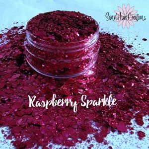 Puede incluir: Un tarro transparente lleno de purpurina color frambuesa, con las palabras "Raspberry Sparkle" en escritura blanca debajo. La purpurina está esparcida alrededor del tarro sobre una superficie blanca. El producto es de SandiMac Creations.