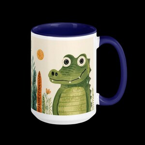 Puede incluir: Taza de cerámica blanca con interior y asa azul marino. La taza presenta una ilustración caprichosa de un caimán verde con ojos grandes, sobre un fondo de plantas, un sol y flores. El diseño es lúdico y colorido.