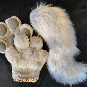 May include: A pair of grey faux fur paw gloves featuring dark grey textured paw pads and a leopard print cuff, accompanied by a long, fluffy grey faux fur tail. The set is designed with soft, shaggy material and detailed paw pad accents.