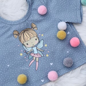 Pode incluir: Uma camiseta azul claro com padrão de bolinhas brancas e um gráfico de fada de desenho animado. A camiseta tem um decote redondo e mangas curtas. A fada está vestindo um vestido rosa e tem cabelo loiro. A camiseta é decorada com pompons coloridos.