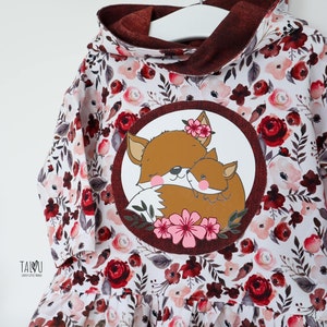 May include: A white hooded dress with a floral print in shades of red, pink, and brown. The dress features a round applique of a mother fox and her cub with pink flowers.