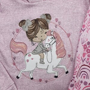 Può includere: Felpa rosa ricamata con una ragazza che cavalca un unicorno. L'unicorno ha una criniera e una coda rosa. La ragazza ha i capelli castani e indossa un vestito grigio. La felpa ha un motivo floreale rosa e bianco sulla manica.
