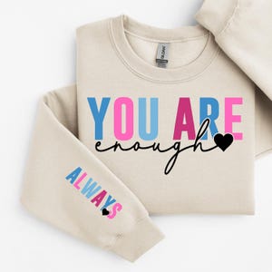 Könnte beinhalten: Ein beiger Pullover mit dem Text "YOU ARE enough" in rosa, blau und schwarzer Schrift mit einem kleinen schwarzen Herz. Der linke Ärmel trägt den Text "ALWAYS" in rosa, blau und schwarzer Schrift mit einem kleinen schwarzen Herz.