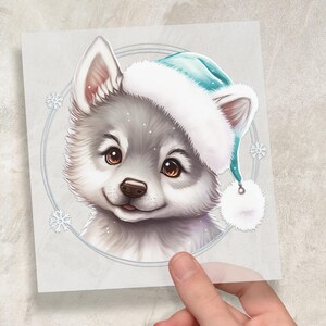 Könnte beinhalten: Eine digitale Illustration eines flauschigen, grauen Welpen mit einer hellblauen und weißen Weihnachtsmütze. Der Welpe hat braune Augen und eine kleine, rosa Zunge. Das Bild ist von einem hellgrauen Kreis mit Schneeflocken umgeben.