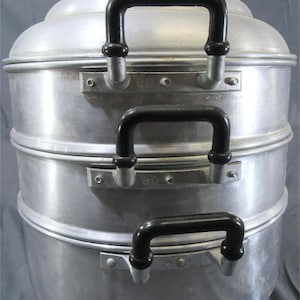 May include: A three-tiered aluminium food steamer with a domed lid. Each section features two black plastic side handles attached with metal brackets. The number 30 is embossed on the metal handle brackets of the middle and top sections.