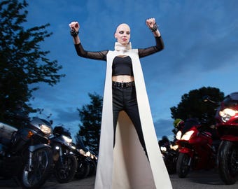 May include: A woman wearing a white cape and black pants stands with her arms raised in front of a row of motorcycles. The woman has short, shaved hair and is wearing white sneakers.