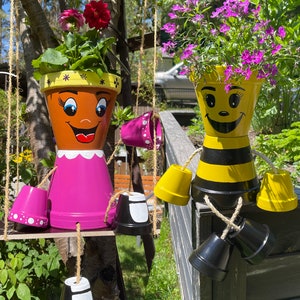 Könnte beinhalten: Zwei dekorative Blumentöpfe aus Terrakotta, bemalt als Mädchen und Biene, hängen im Freien. Der Mädchen-Topf ist braun mit einem rosa Kleid, der Bienen-Topf ist gelb und schwarz. Beide Töpfe enthalten bunte Blumen.