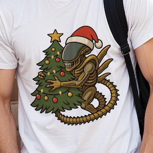 Könnte beinhalten: Weißes T-Shirt mit einem Cartoon-Alien, das einen Weihnachtsmannhut trägt und einen Weihnachtsbaum umarmt. Das Alien ist gold- und braunfarben, und der Baum ist grün mit roten und goldenen Ornamenten. Ein schwarzer Rucksack ist rechts zu sehen.