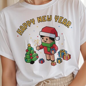 Puede incluir: Camiseta blanca con un gráfico pixelado que dice "HAPPY NEW YEAR". El gráfico presenta un personaje pixelado con un gorro de Papá Noel, bufanda y sosteniendo una bengala, con un árbol de Navidad y regalos envueltos.