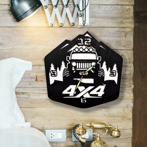 Op de afbeelding: Zwart-witte wandklok met een 4x4 off-road voertuig ontwerp. De klok heeft een berg silhouet, bomen en de tekst "4x4". De klok is gemonteerd op een houten muur.