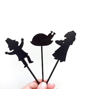 May include: Three black silhouette cake toppers featuring a person in a hat, a roasted turkey on a platter, and a person holding a dish. Each figure is attached to a thin black stick, designed for insertion into food items for decoration.