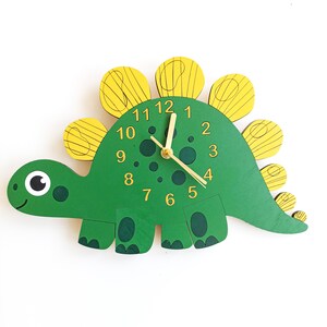 May include: A green dinosaur-shaped wall clock with yellow accents. The clock features a smiling face, a round eye, and gold-colored clock hands. The numbers are in a gold color. The clock is made of wood.