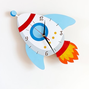 Könnte beinhalten: Eine weiße raketenförmige Uhr mit blauen und roten Akzenten. Das Zifferblatt der Uhr hat schwarze Zeiger und schwarze Zahlen. Die Rakete hat eine gelbe und orangefarbene Flamme.