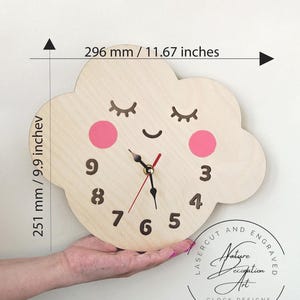 May include: A light-colored wooden wall clock shaped like a cloud with a smiling face, pink cheeks, and dark brown numbers. The clock measures 11.67 inches wide by 9.9 inches high. It features black hour and minute hands and a red second hand.
