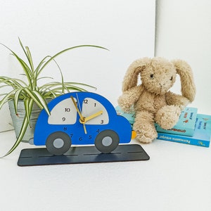 May include: A blue car-shaped clock with a white clock face and gold hands sits on a black base. A plush bunny and a potted plant are in the background. The clock has the numbers 1-12.