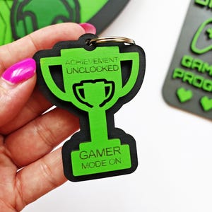 May include: A black and green trophy-shaped keychain with the words "ACHIEVEMENT UNLOCKED" and "GAMER MODE ON" in white. The keychain has a gold ring for attaching to keys. The design is a gaming theme.