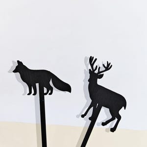 Peut inclure: Deux piquets de jardin noirs en forme de silhouettes. L'un représente un renard, l'autre un cerf avec des bois. Les piquets sont placés sur un fond blanc et beige. Ces piquets décoratifs ajoutent une touche de nature à tout jardin.