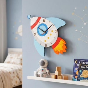 Op de afbeelding: Een raketvormige wandklok in wit, rood en blauw, met zwarte wijzers. De klok is bevestigd aan een lichtblauwe muur, met daaronder een plank met een astronautenfiguur, houten blokken en ruimteboeken. De klok meet ongeveer 30 cm in hoogte.