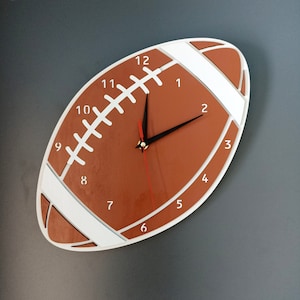 Könnte beinhalten: Eine braun-weiße Wanduhr in Football-Form. Die Uhr hat schwarze Zeiger und einen roten Sekundenzeiger. Weiße Zahlen markieren die Stunden. Das Design der Uhr ahmt das Aussehen eines Footballs mit weißen Streifen und Nähten nach.