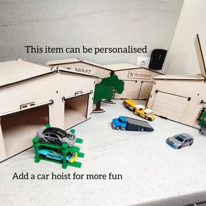 Könnte beinhalten: Ein hölzernes Spielzeug-Garagenset mit zwei Garagentoren, einer Autohebebühne und einem kleinen Baum. Die Garagentore sind mit "Mikey" und "Brelsford" beschriftet. Vor der Garage stehen mehrere Spielzeugautos, darunter ein blauer Autotransporter, ein gelber Kipplaster und ein silbernes Auto.