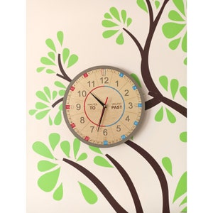 Peut inclure: Une horloge ronde en bois avec une bordure grise, présentant un cadran en bois clair. L'horloge a des aiguilles noires et affiche les mots "Minutes To" et "Minutes Past". Les chiffres sont en noir et rouge, avec une section bleue pour "Past".