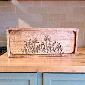 May include: A rectangular wooden serving tray with rounded edges. The tray features a detailed etched design of mushrooms and foliage. The wood has a natural grain and a warm, light brown color, suitable for serving food.