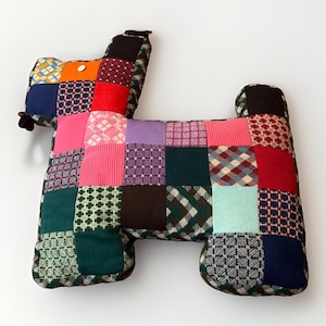 May include: A stuffed dog toy constructed from a patchwork of various fabric squares in diverse colors and patterns, including red, pink, green, blue, purple, and orange. The toy features a small white button eye and a dark brown fabric ear.