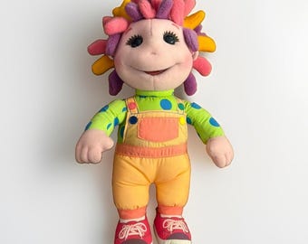 May include: A plush doll with a light pink face and dark eyes, featuring a vibrant hairstyle of multi-colored fabric loops in yellow, orange, pink, and purple. The doll wears a lime green long-sleeve shirt with blue polka dots, orange overalls, and red sneakers.