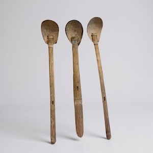 May include: Three antique wooden spoons with elongated handles and oval-shaped bowls. The spoons have a light brown color with visible wood grain and appear to be handmade. The bowls have a slightly darker hue, suggesting a different material or age.