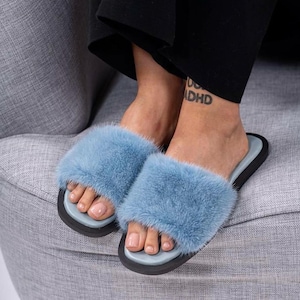 May include: A pair of light blue fur slides with a black sole and light blue footbed. The slides are on a gray upholstered chair. The image shows a close-up of the slides.