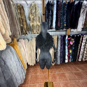May include: A black fur collar with a hood on a black mannequin. The mannequin is standing on a gold stand. The background is a wall with many fur coats hanging on racks.