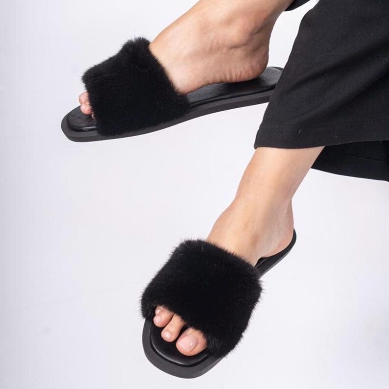 May include: Black faux fur slide sandals. The sandals have a wide strap of soft, fluffy material across the top of the foot. The soles are black and flat. The image shows the sandals being worn.