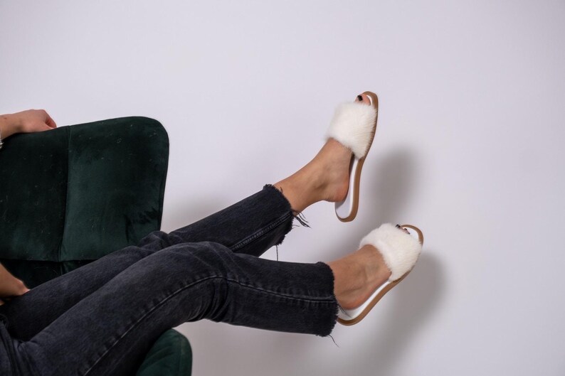 May include: White fluffy slide sandals with a brown sole are worn with black denim jeans. The sandals have a square toe and a soft, plush upper. The jeans are distressed at the ankles. The person is sitting in a dark green velvet chair.