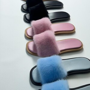 May include: A variety of fur-covered slide sandals in a row. Colors include beige, white, black, pink, and blue. Each sandal has a flat sole and a fluffy fur strap. The soles are approximately 1 inch thick.