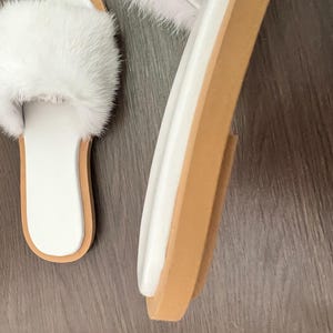 May include: White faux fur slide sandals with a white footbed and a light brown sole. The sandals have a fluffy strap across the top. The sole is approximately 1 inch thick. The sandals are shown on a dark brown wooden surface.