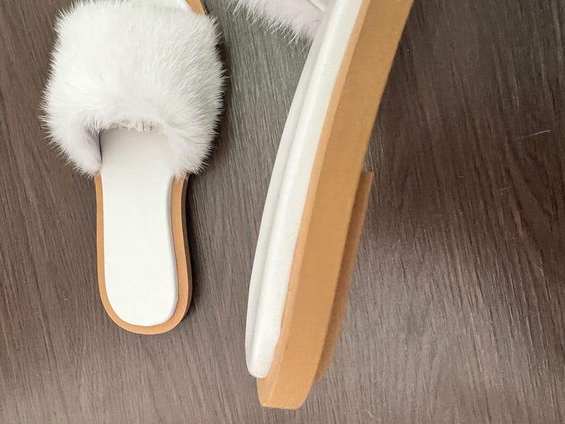 May include: White faux fur slide sandals with a white footbed and a light brown sole. The sandals have a fluffy strap across the top. The sole is approximately 1 inch thick. The sandals are shown on a dark brown wooden surface.