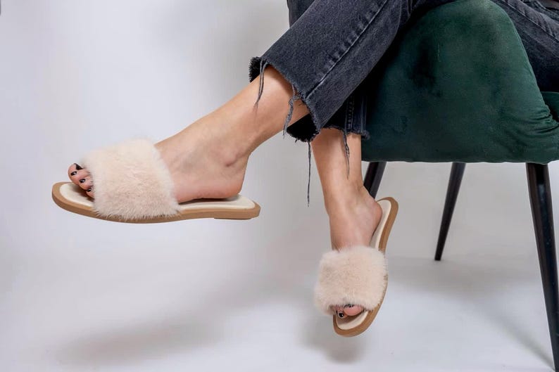 May include: A pair of beige faux fur slippers with a light brown sole. The slippers are being worn by a person sitting in a green chair. The person is wearing black jeans.